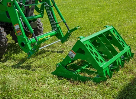 Right side view of an MG20F root grapple lying in the grass in front of a loader with quick-attach capabilities