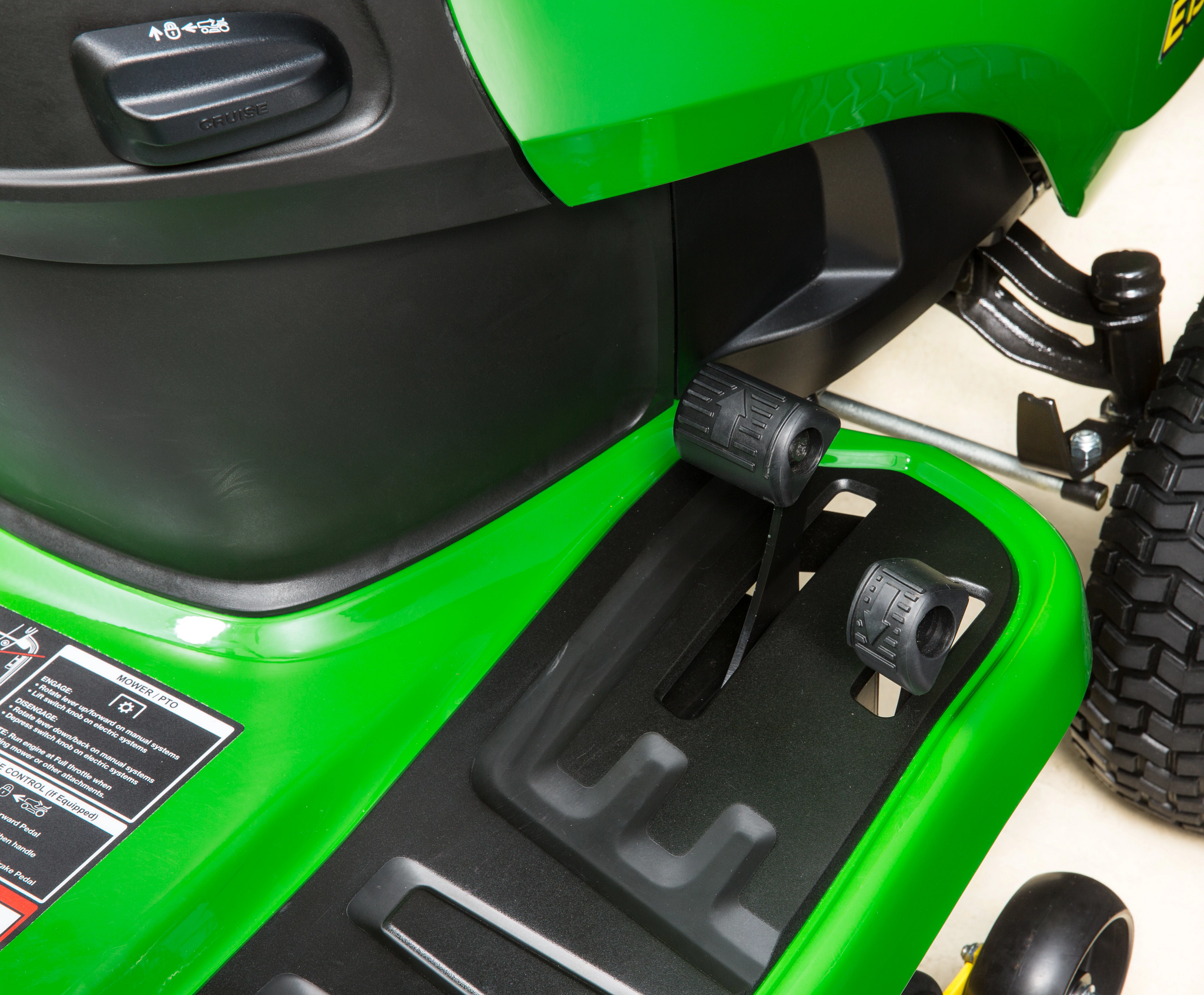 Close-up of a riding mower footwell showing forward and reverse pedals, black textured floor panel, and adjacent control housing.