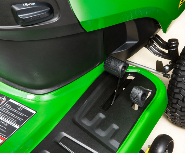 Close-up of a riding mower footwell showing forward and reverse pedals, black textured floor panel, and adjacent control housing.