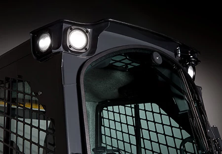 Close-up of the upper cab of a compact loader, showing bright LED work lights mounted on the roof and protective metal mesh around the cab