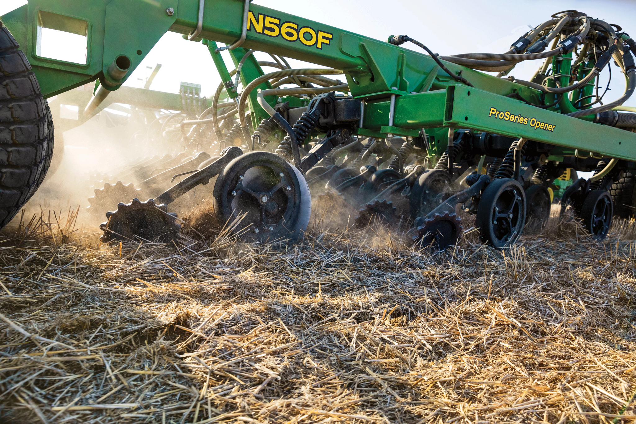 Green John Deere N560F Air Drill operating in a field covered with crop residue, with multiple row units and discs churning soil and kicking up dust