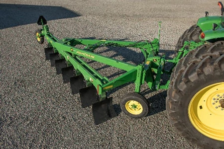 Angled side view of a 955 moldboard plow's green V-style mainframe on gravel, with two tractor wheels in the right side of the frame
