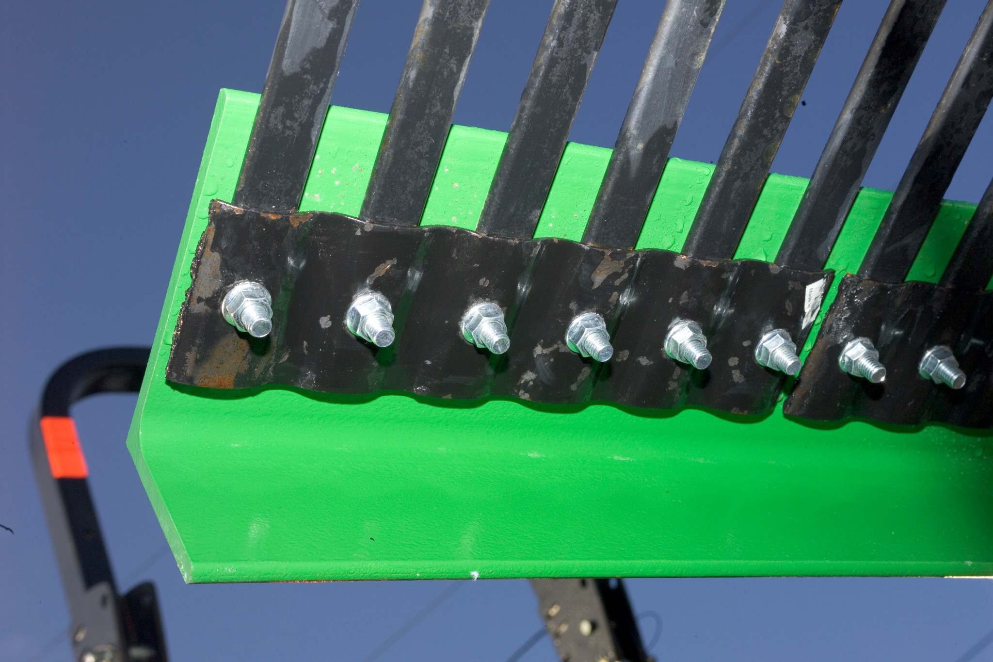 Close-up view of a green metal attachment edge with several black vertical tines bolted along the top using silver hex bolts, shown against a blue sky