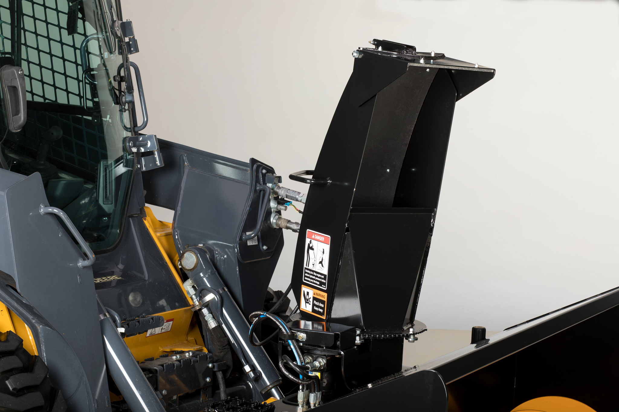 A close-up of a construction snowblower attachment with a discharge chute connected by hydraulic arms, shown with visible hoses and warning labels