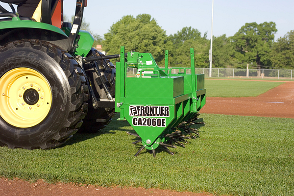 Green CA2060E Core Aerator attached to a tractor, shown operating on grass with spiked coring wheels engaged near the edge of a baseball infield