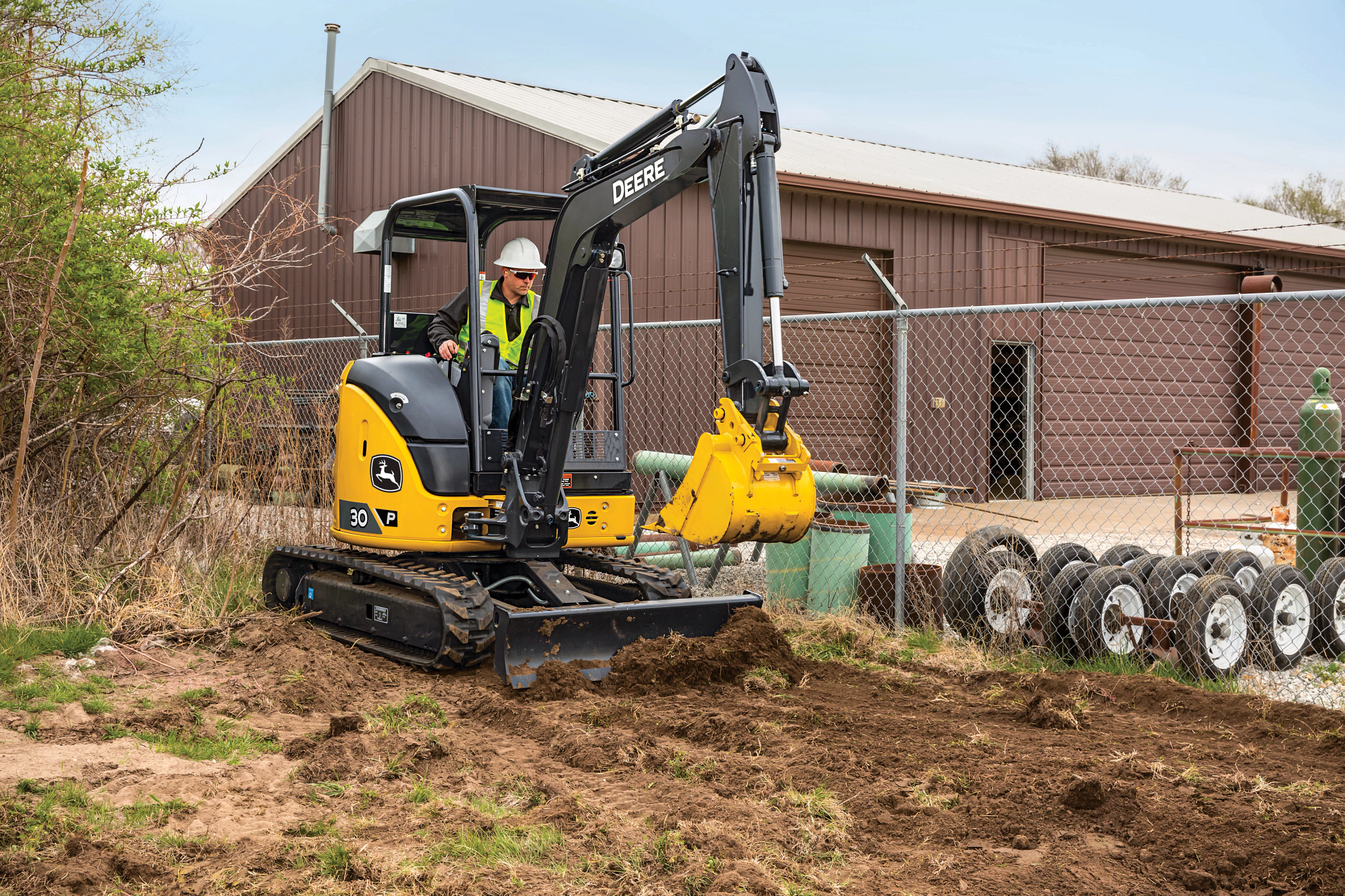 Worker operating a compact John Deere excavator, digging soil at a fenced work site