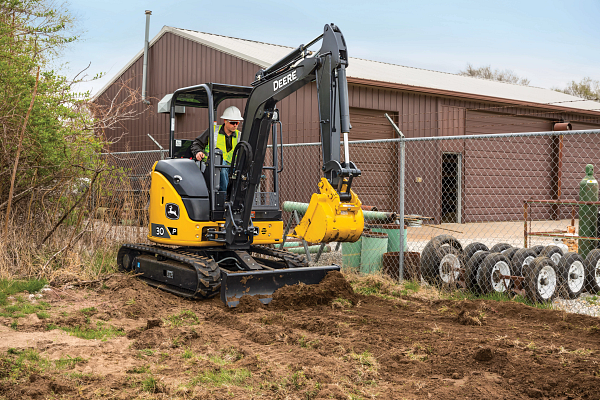 Worker operating a compact John Deere excavator, digging soil at a fenced work site