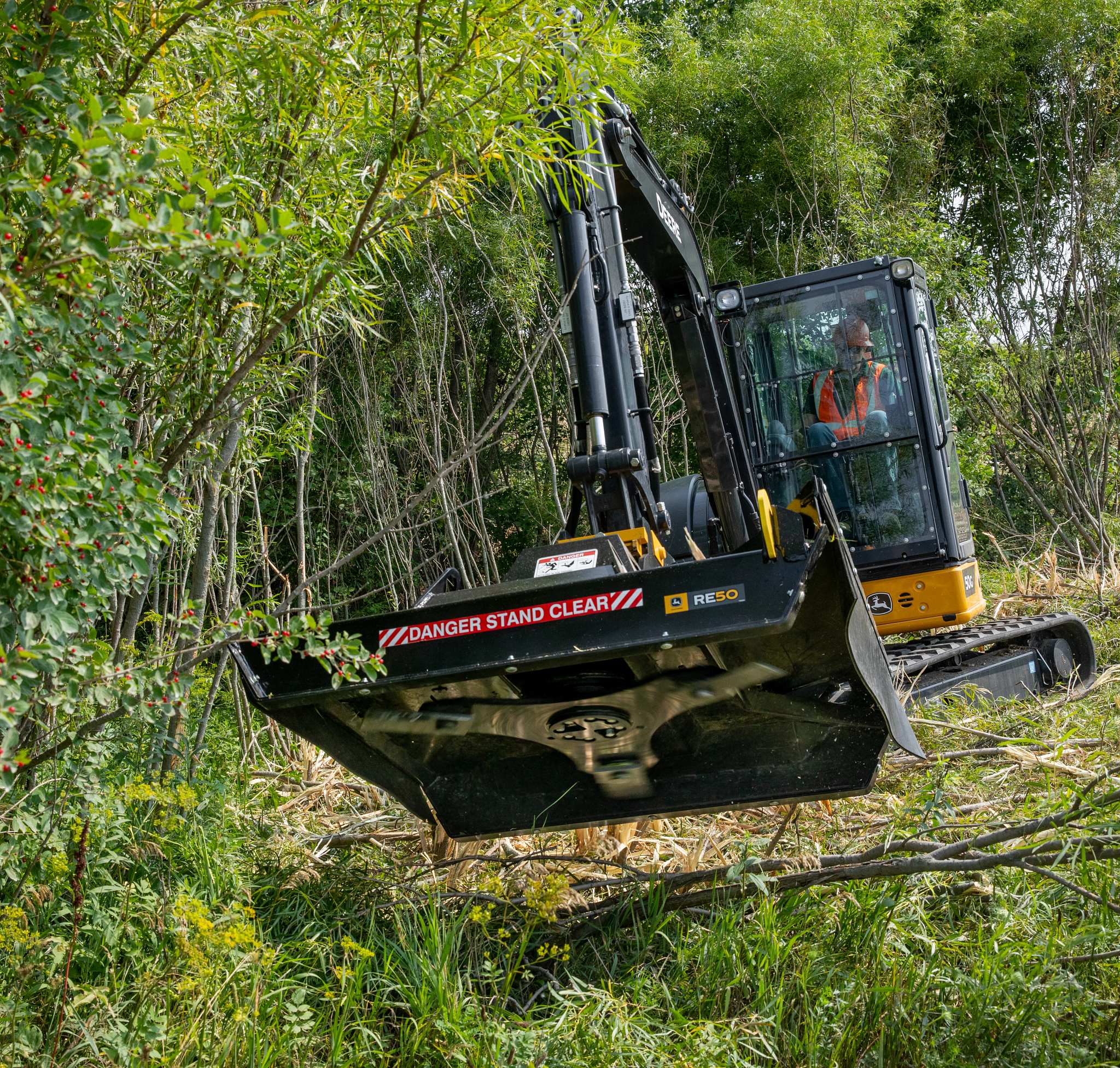 Front view of an RE50 brush cutter hoisted above shrubs and sticks in the woods by its attached compact excavator.