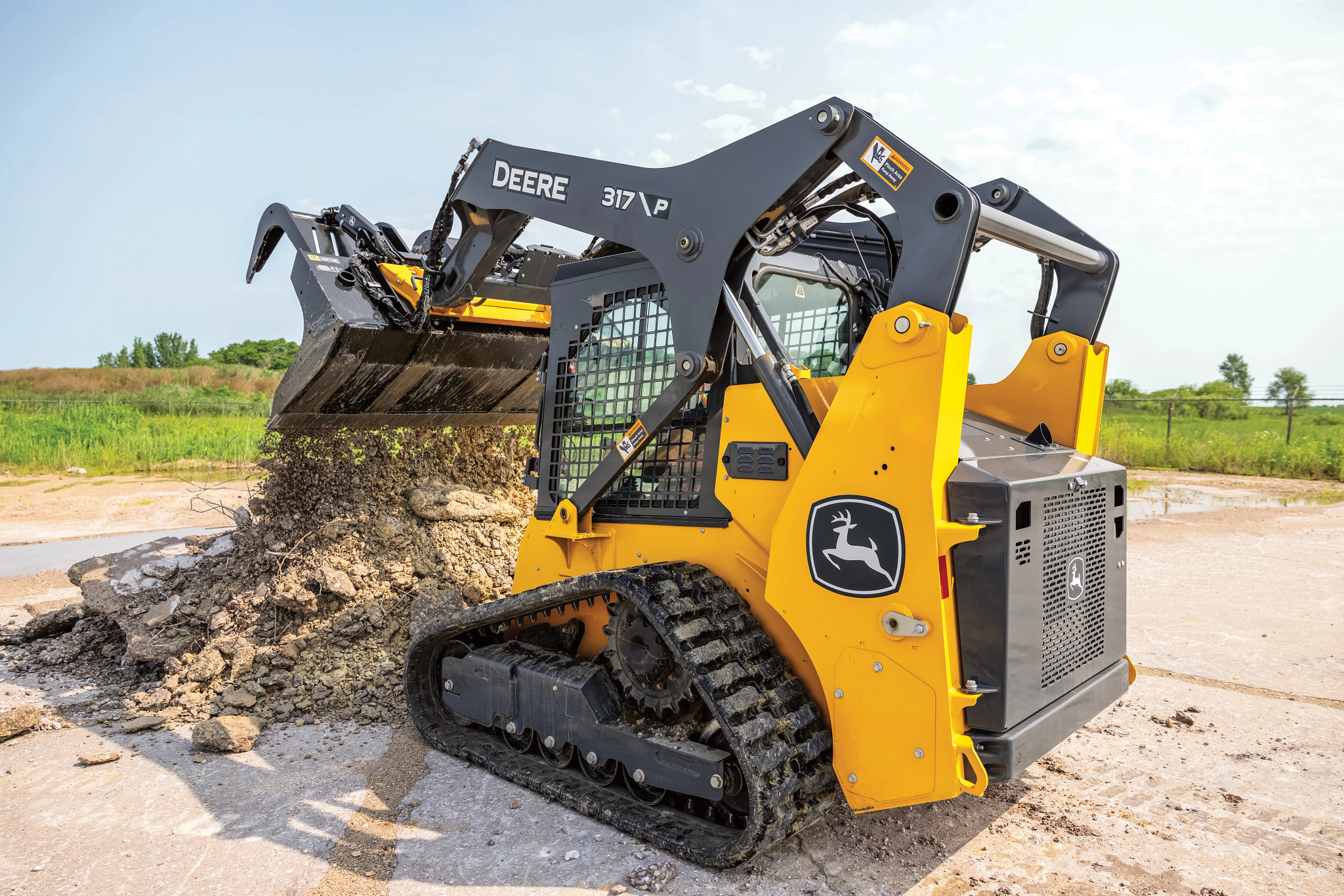Yellow and black John Deere 317G compact track loader with rubber tracks, lifting and dumping a bucket of dirt and gravel onto a construction site surface, with a grassy field and trees in the background under a clear sky.