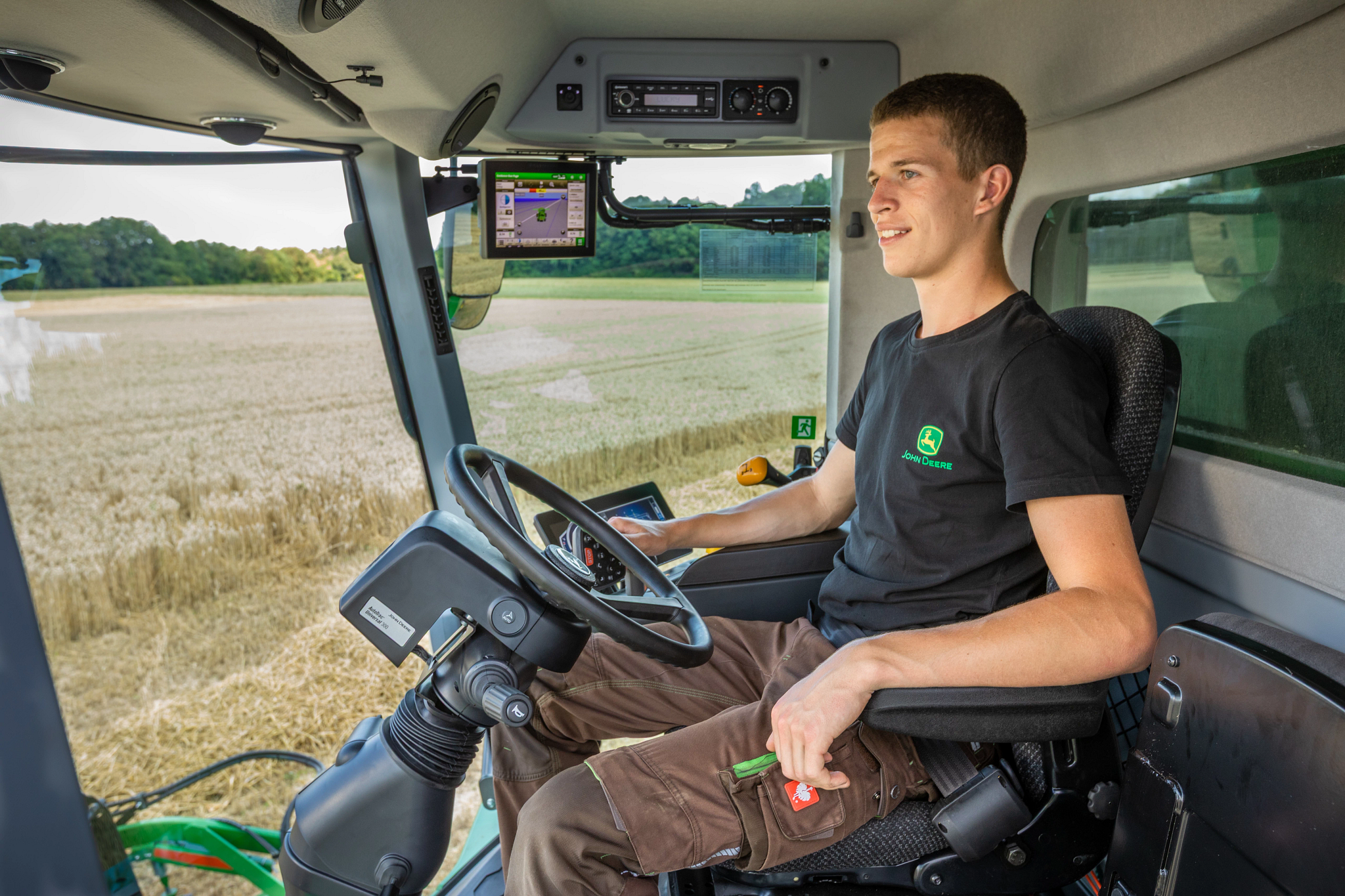 A modern agricultural machine cab, operating controls while viewing a field through large windows, with a mounted display screen visible above the steering wheel.