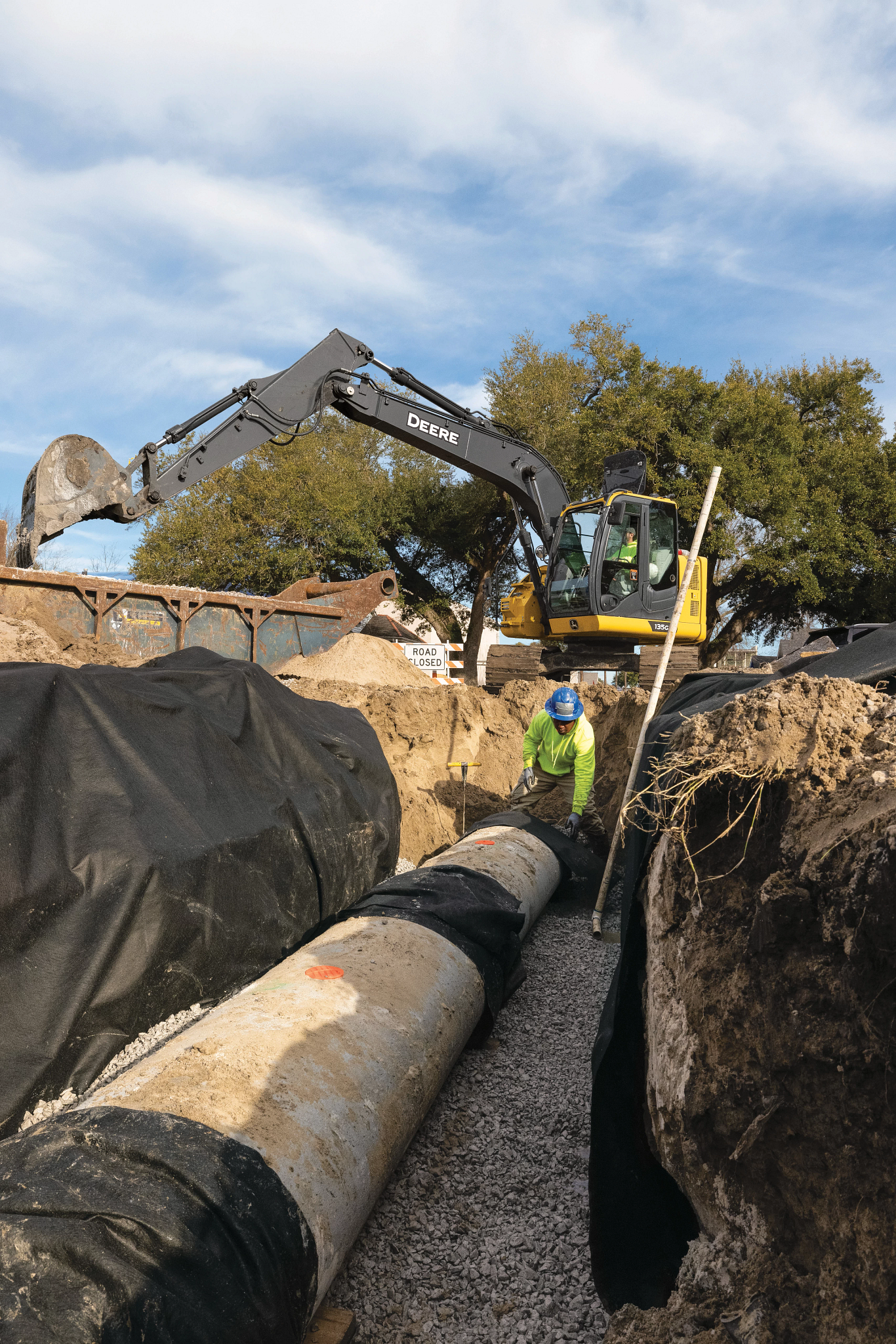 Construction worker in a trench beside a large pipeline while an excavator extends its arm overhead with green foliage and a partly cloudy sky appear in the background