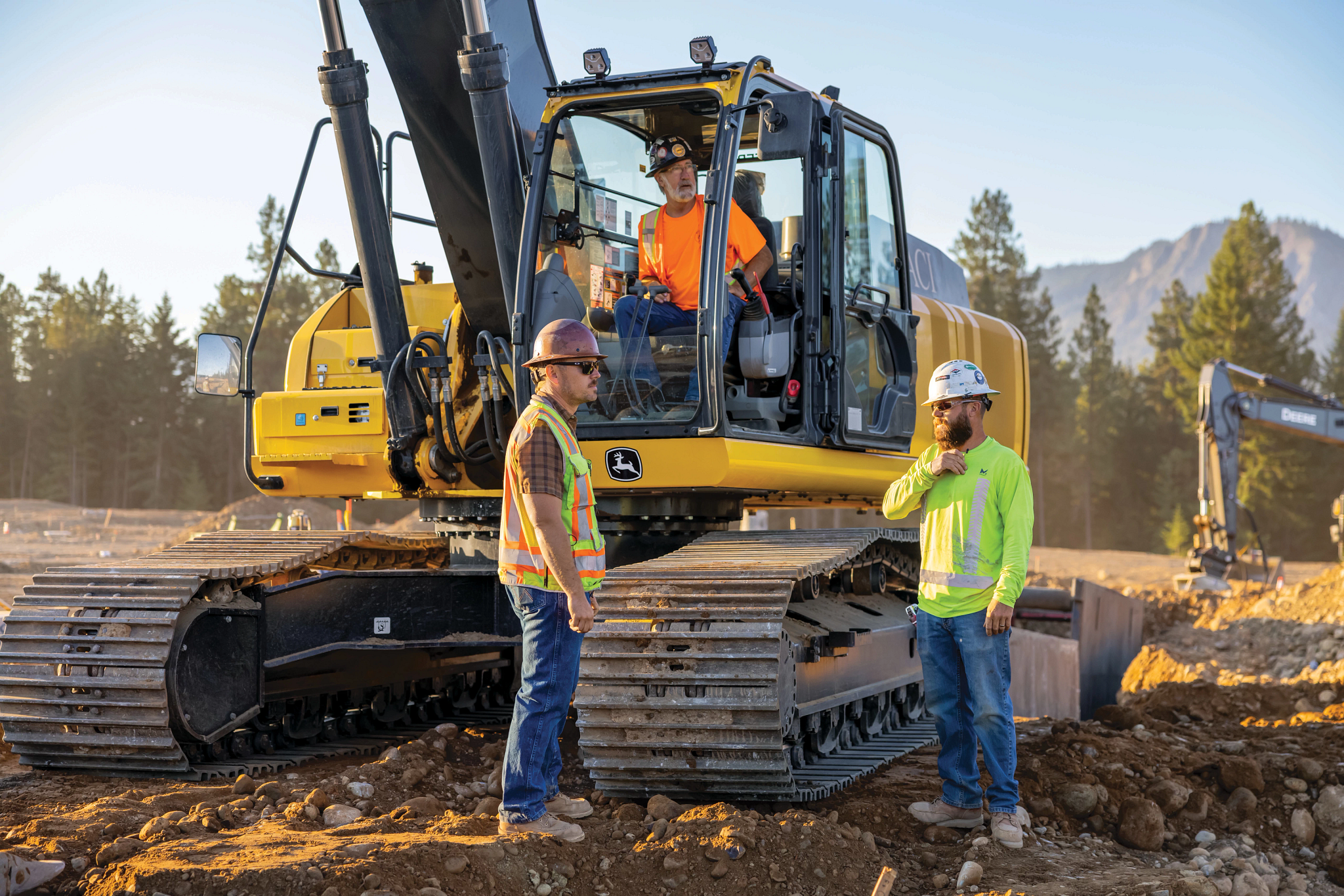 Two construction workers standing beside a large yellow excavator with metal tracks, while a third worker sits inside the enclosed cab