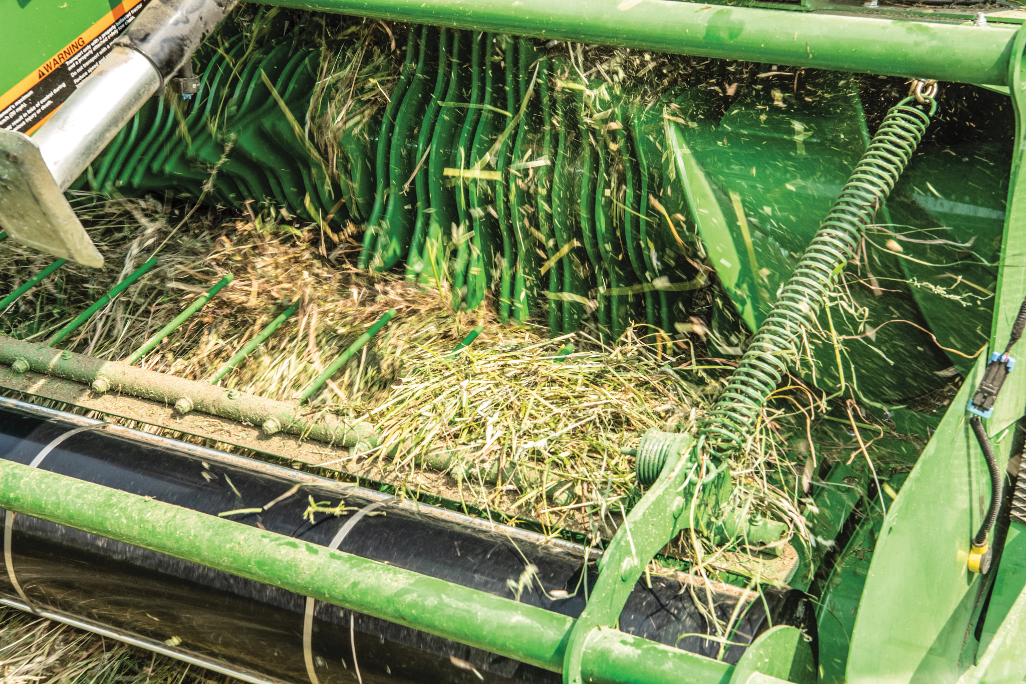 Close-up of a John Deere baler pickup showing rotating green tine bars and feed rollers lifting and guiding cut hay into the baling chamber.