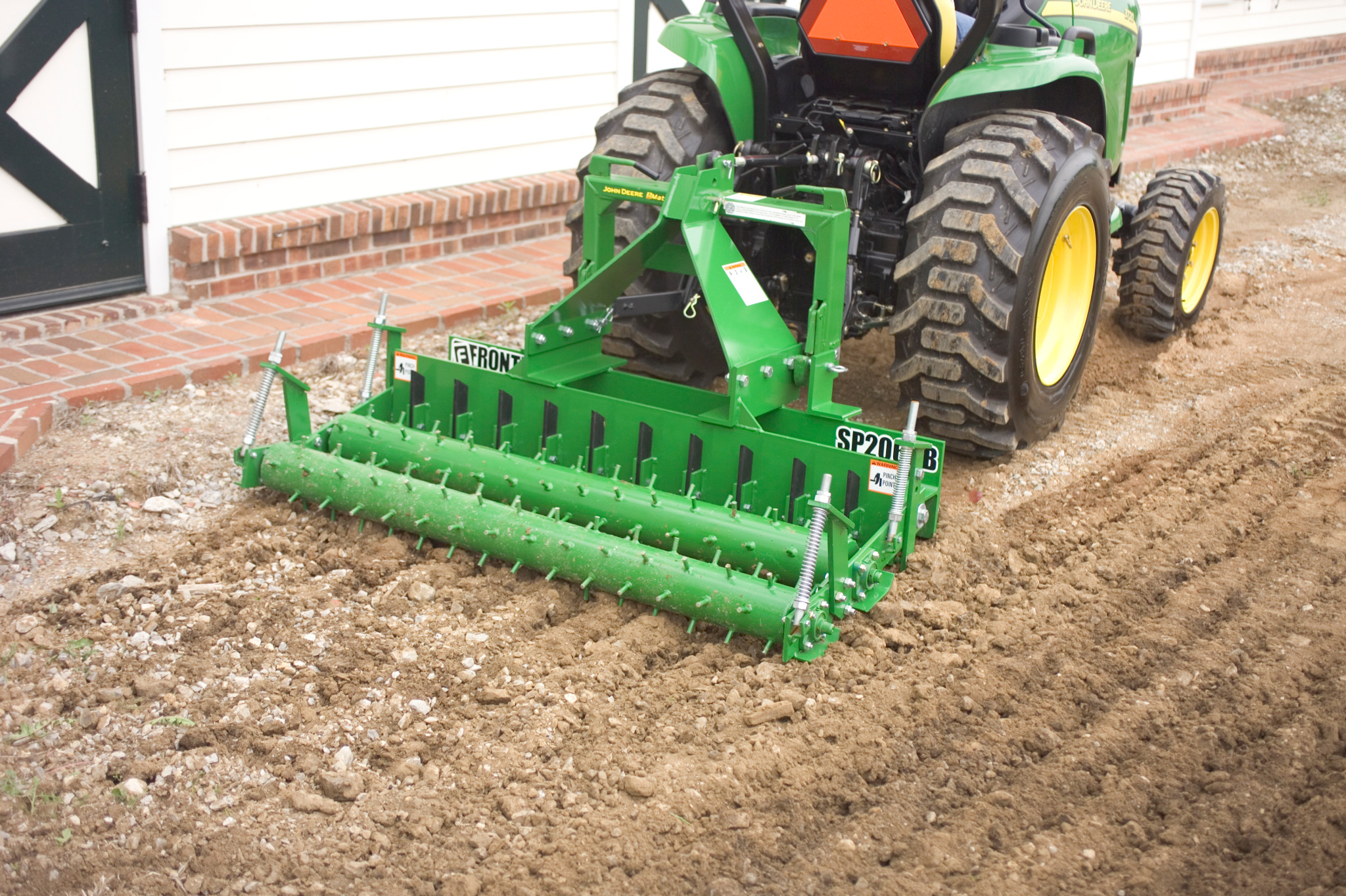 Compact tractor pulling a green soil pulverizer over a dirt surface, leaving a leveled and finely textured seedbed behind it