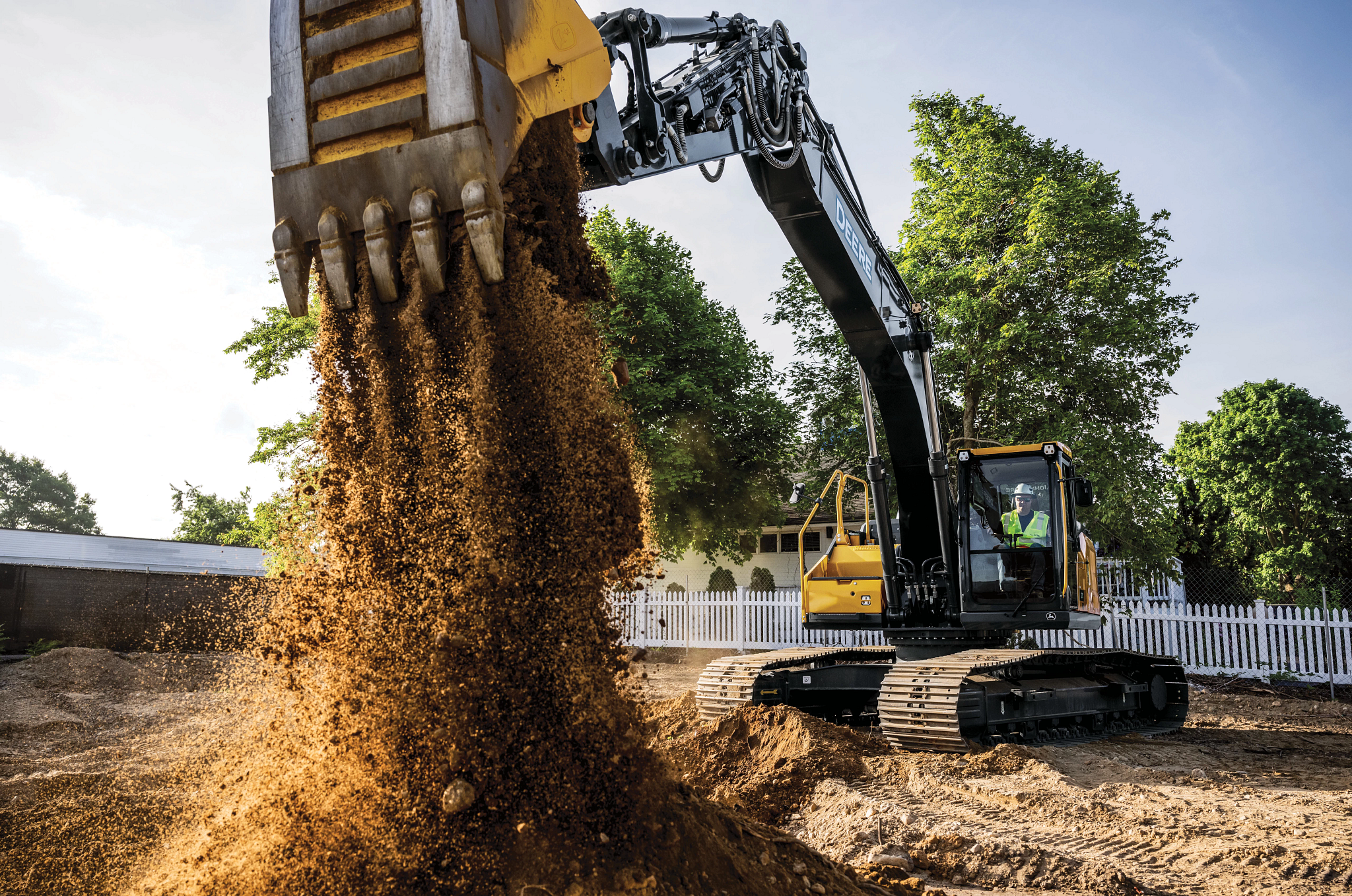 A construction excavator with a large metal bucket pours a cascade of brown soil onto the ground, creating a growing dirt pile