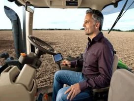 Inside view of a tractor cab showing an operator seated at the controls, with hands near the steering wheel and touchscreen display.