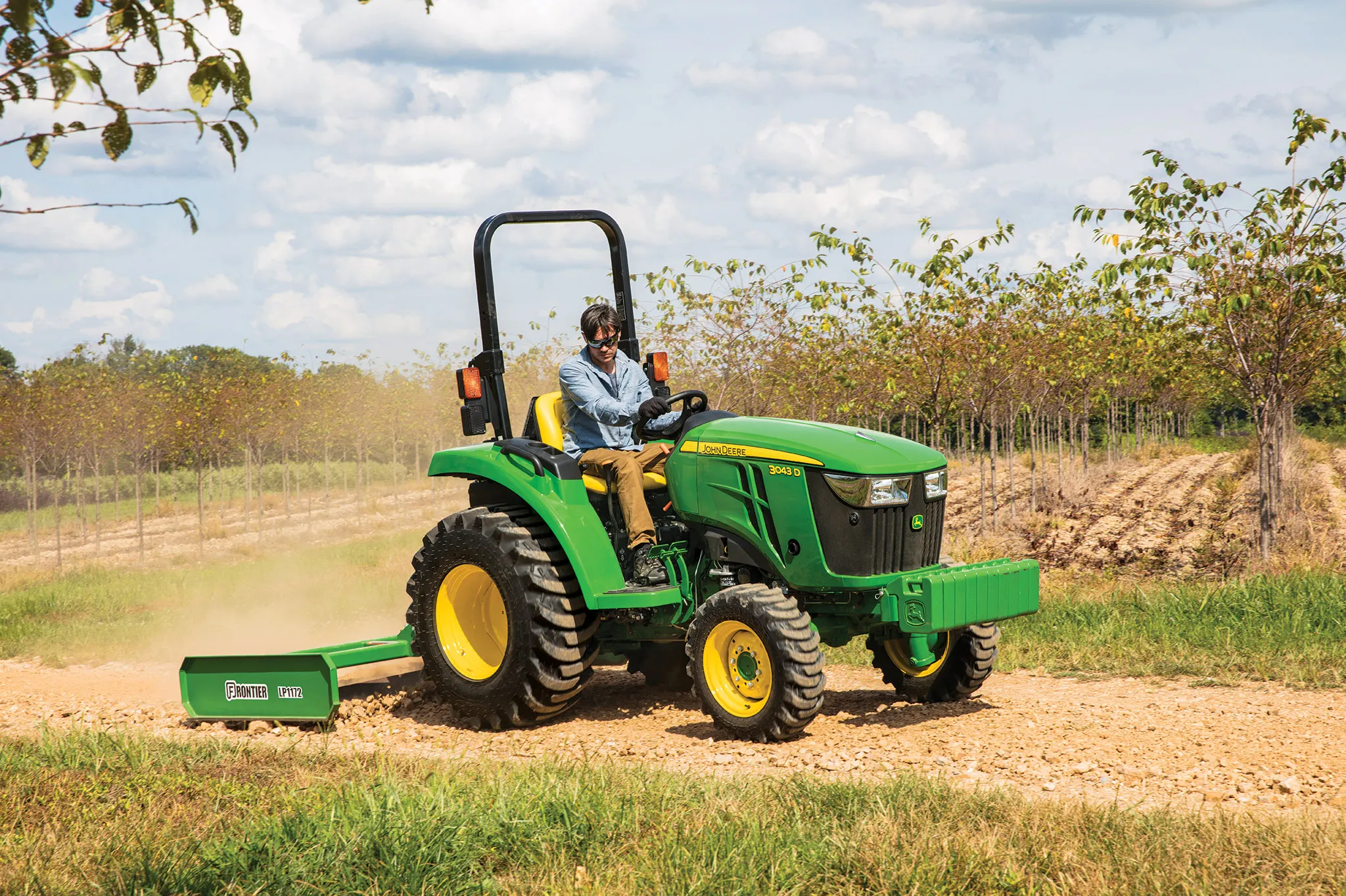 A person drives a green tractor pulling a land plane attachment along a dirt path beside rows of trees under a partly cloudy sky