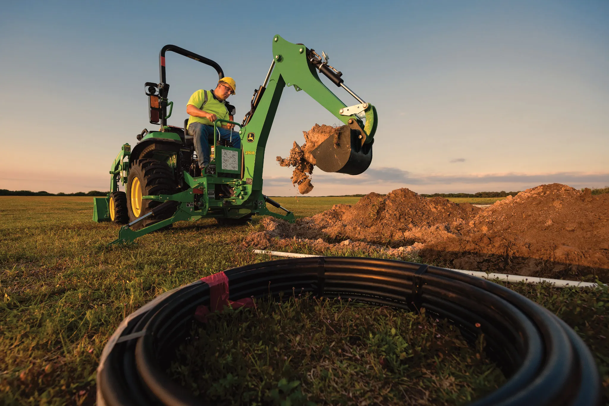 Green compact tractor with a front loader scoops dirt near a circular stack of black pipes in a grassy field at sunset, with a soil mound nearby and warm evening light under a clear sky