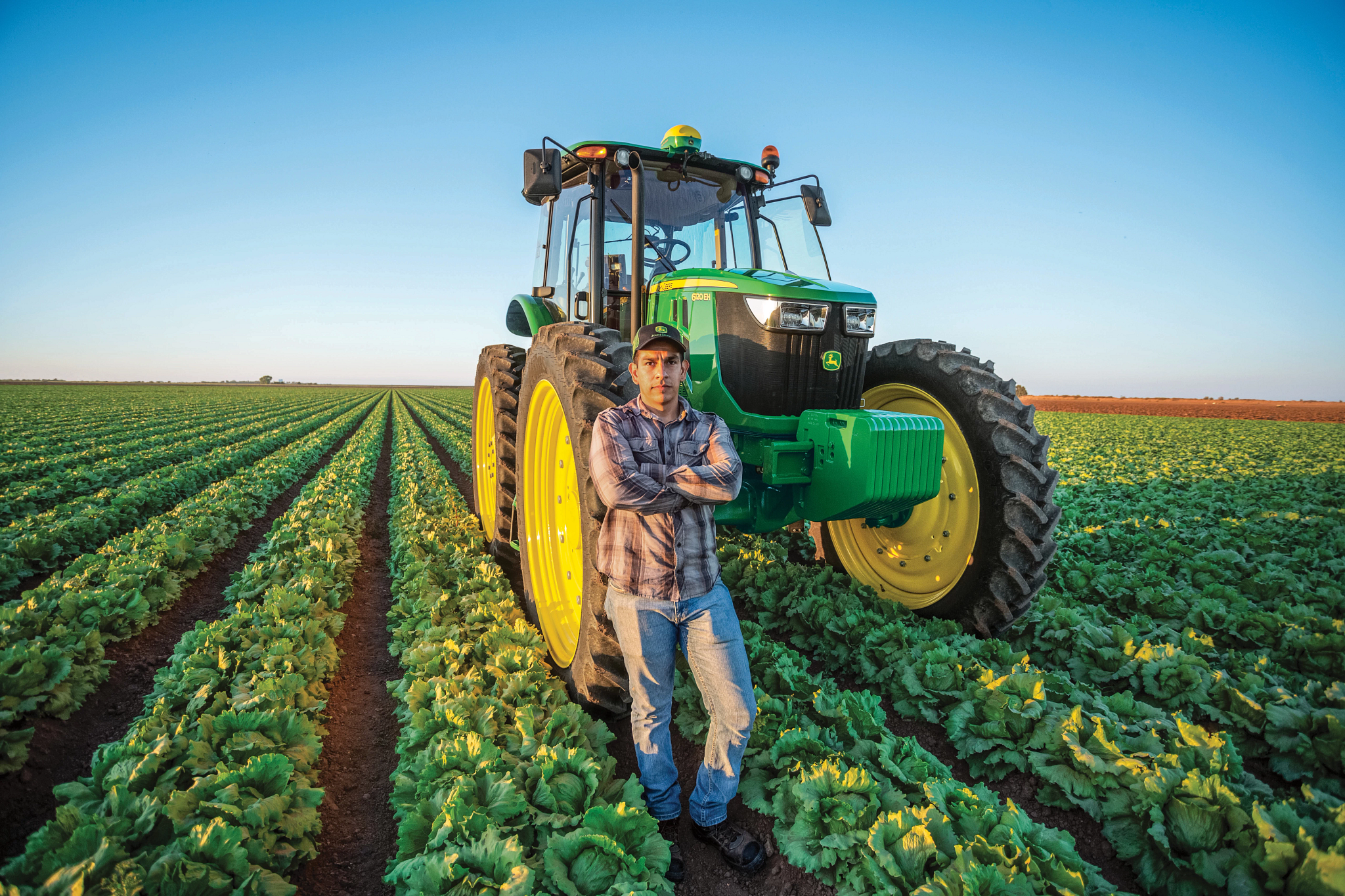 John Deere row‑crop tractor parked between rows of leafy green crops, with large yellow wheels and enclosed cab; a person stands leaning against the front tire under a clear blue sky.