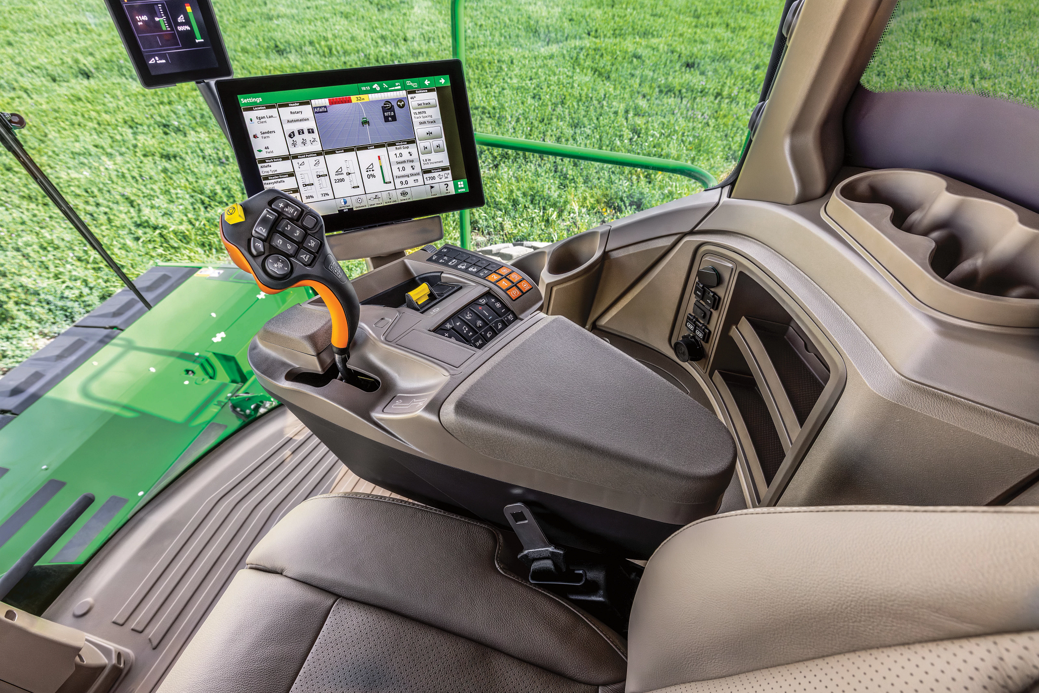 Interior of a John Deere self‑propelled windrower cab showing operator seat, armrest‑mounted joystick, touchscreen display, and control panel, with green crop visible through the front window.
