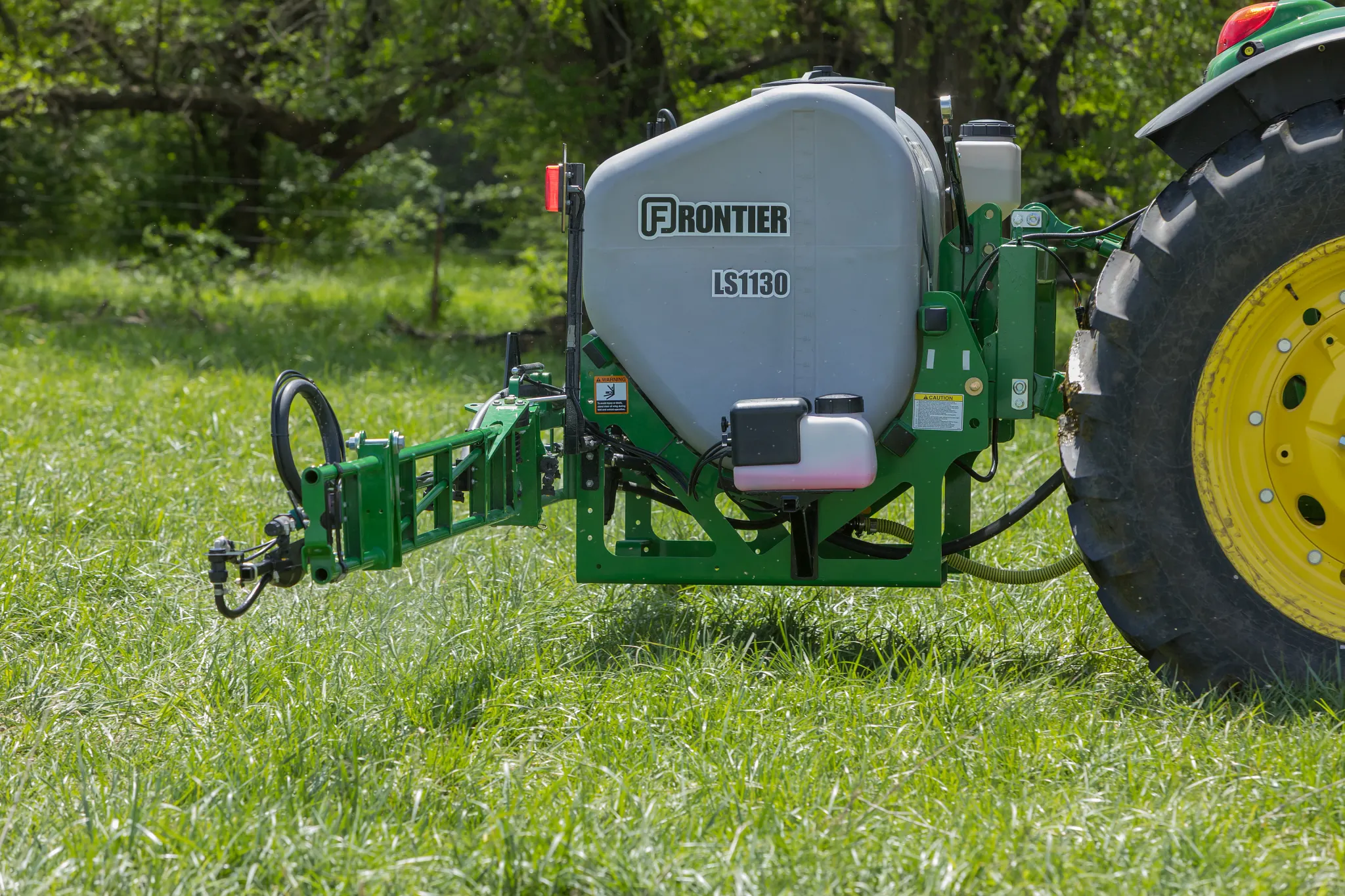Frontier LS1130 tractor-mounted sprayer with a gray tank spraying attachment positioned on grass, connected to a tractor tire, outdoors in a grassy field with trees in the background