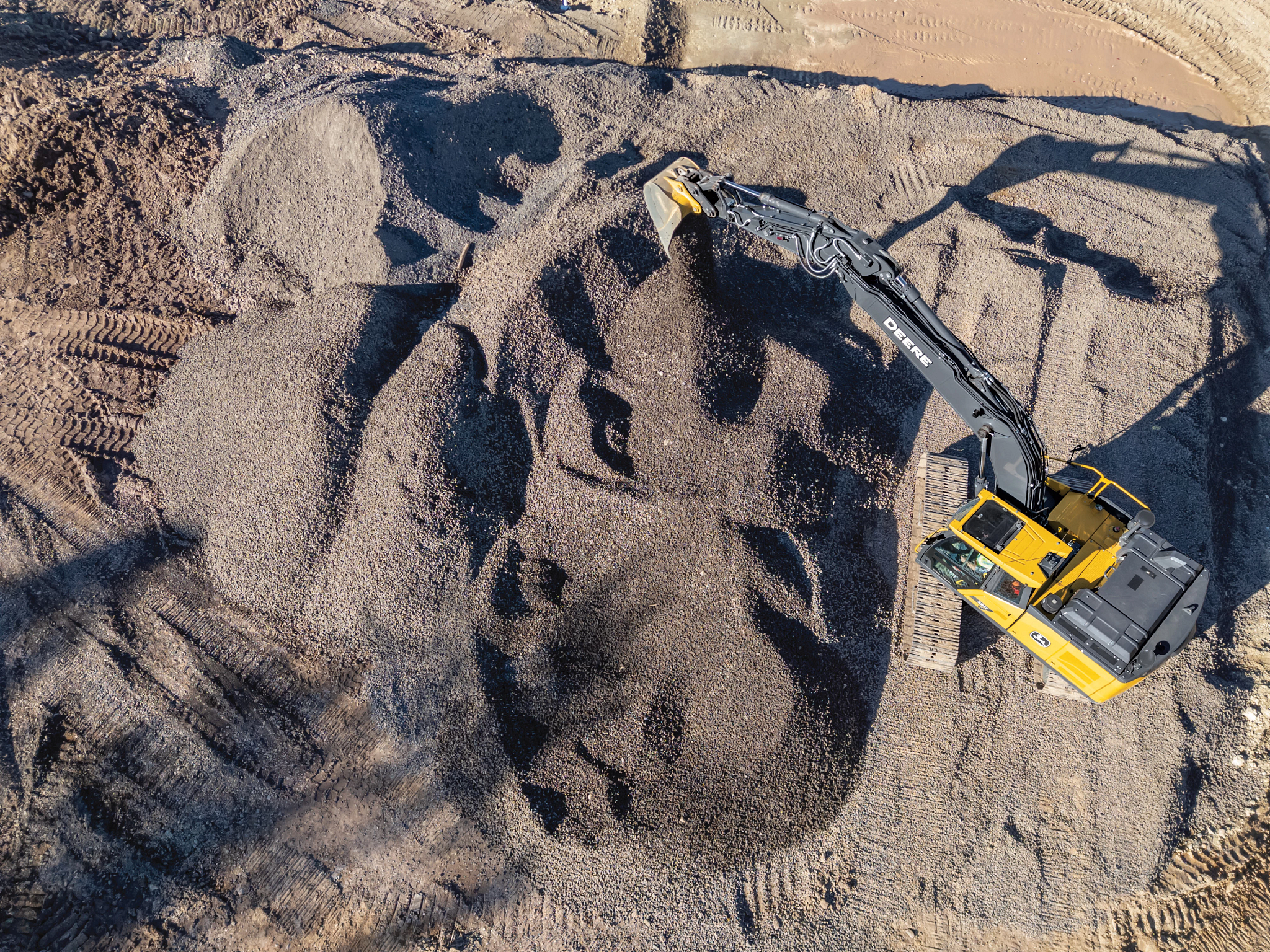 Aerial view of a yellow excavator positioned on a large gravel site, extending its arm to pour a stream of small stones onto a mound below