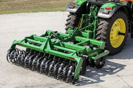 A green tractor towing a rear-mounted tillage attachment with multiple rows of black metal rollers and discs, positioned on a concrete surface