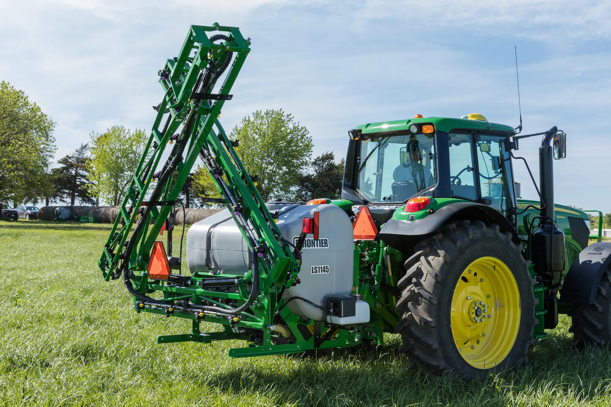 Agricultural tractor with yellow wheels parked on a grassy field, towing a folded boom sprayer with metal arms raised vertically and a large tank mounted behind the tractor under a clear blue sky.