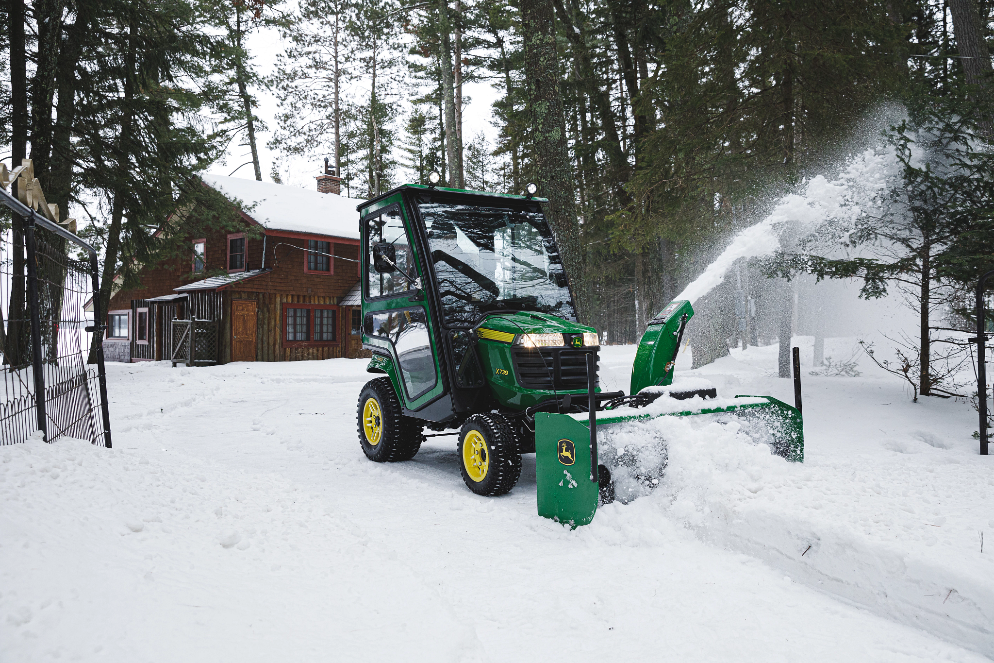 A compact tractor with an enclosed cab using a front-mounted snow blower to clear deep snow, throwing snow to the side.