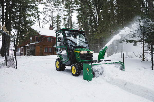 A compact tractor with an enclosed cab using a front-mounted snow blower to clear deep snow, throwing snow to the side.