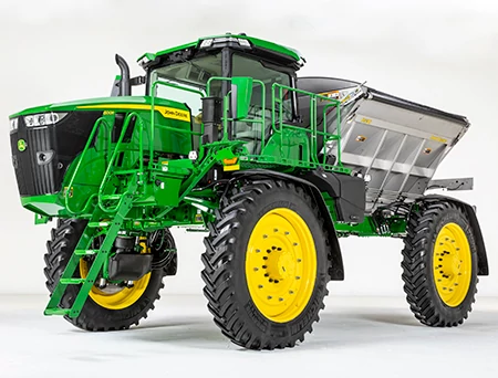 A large spreader with yellow wheels, an elevated cab, access ladder, tall chassis, and a large metal hopper mounted on the rear, displayed against a white background
