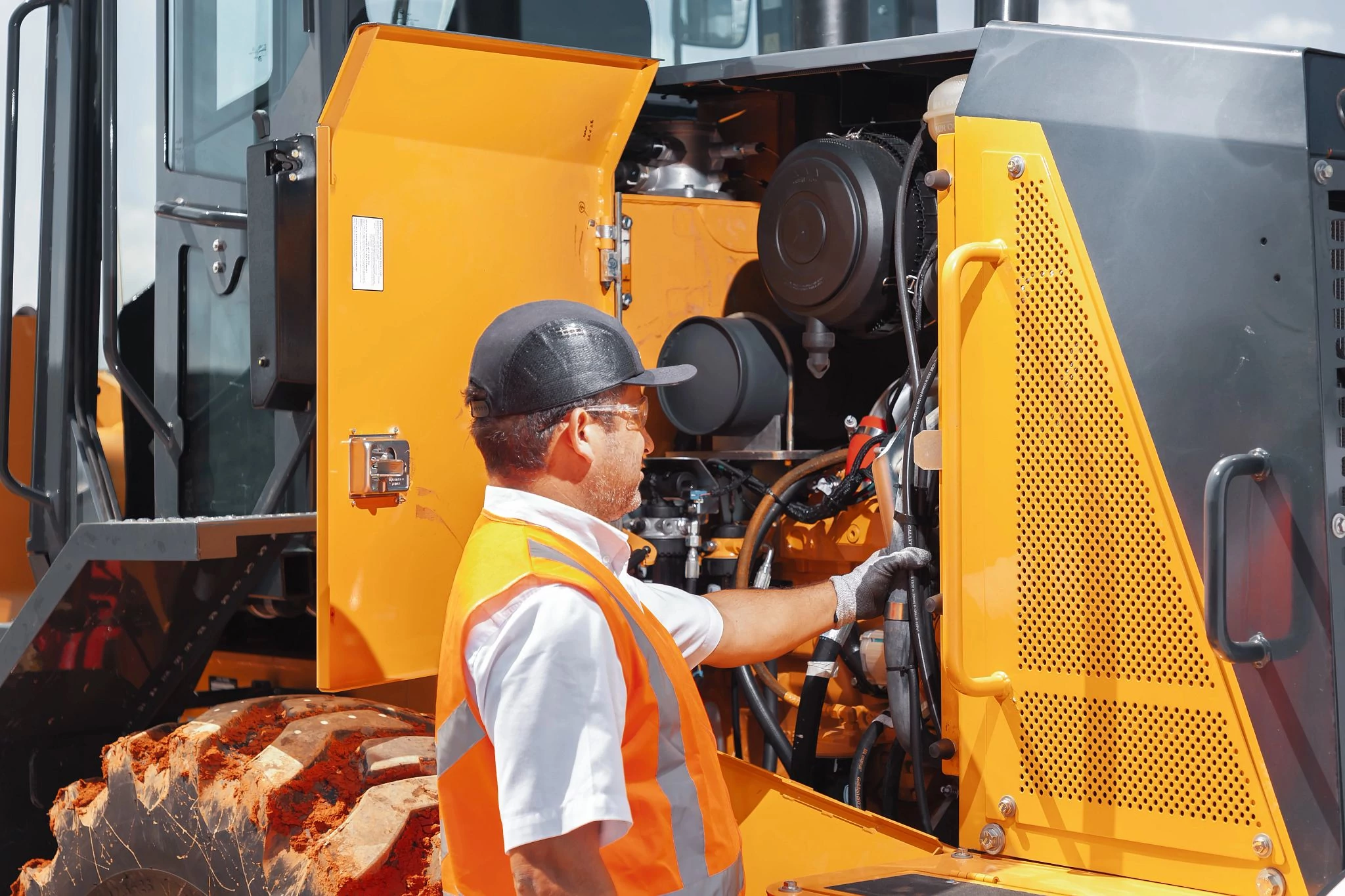 A person wearing a hard hat and high-visibility orange safety vest stands beside wheel loader with an open engine compartment, inspecting internal mechanical components.