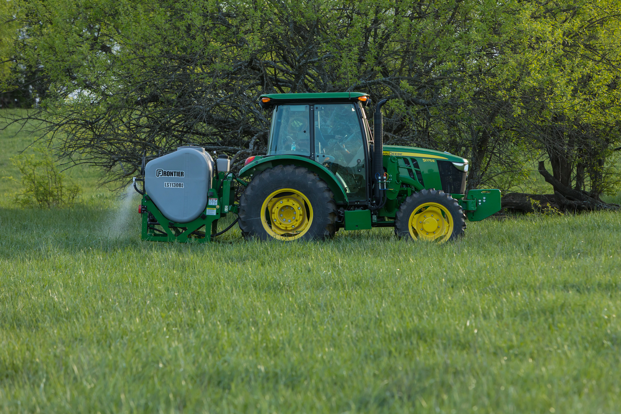 Tractor with yellow wheels pulling a tank sprayer, parked in a grassy field with leafy trees in the background on a sunny day.
