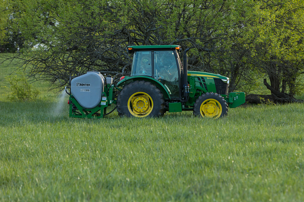 Tractor with yellow wheels pulling a tank sprayer, parked in a grassy field with leafy trees in the background on a sunny day.