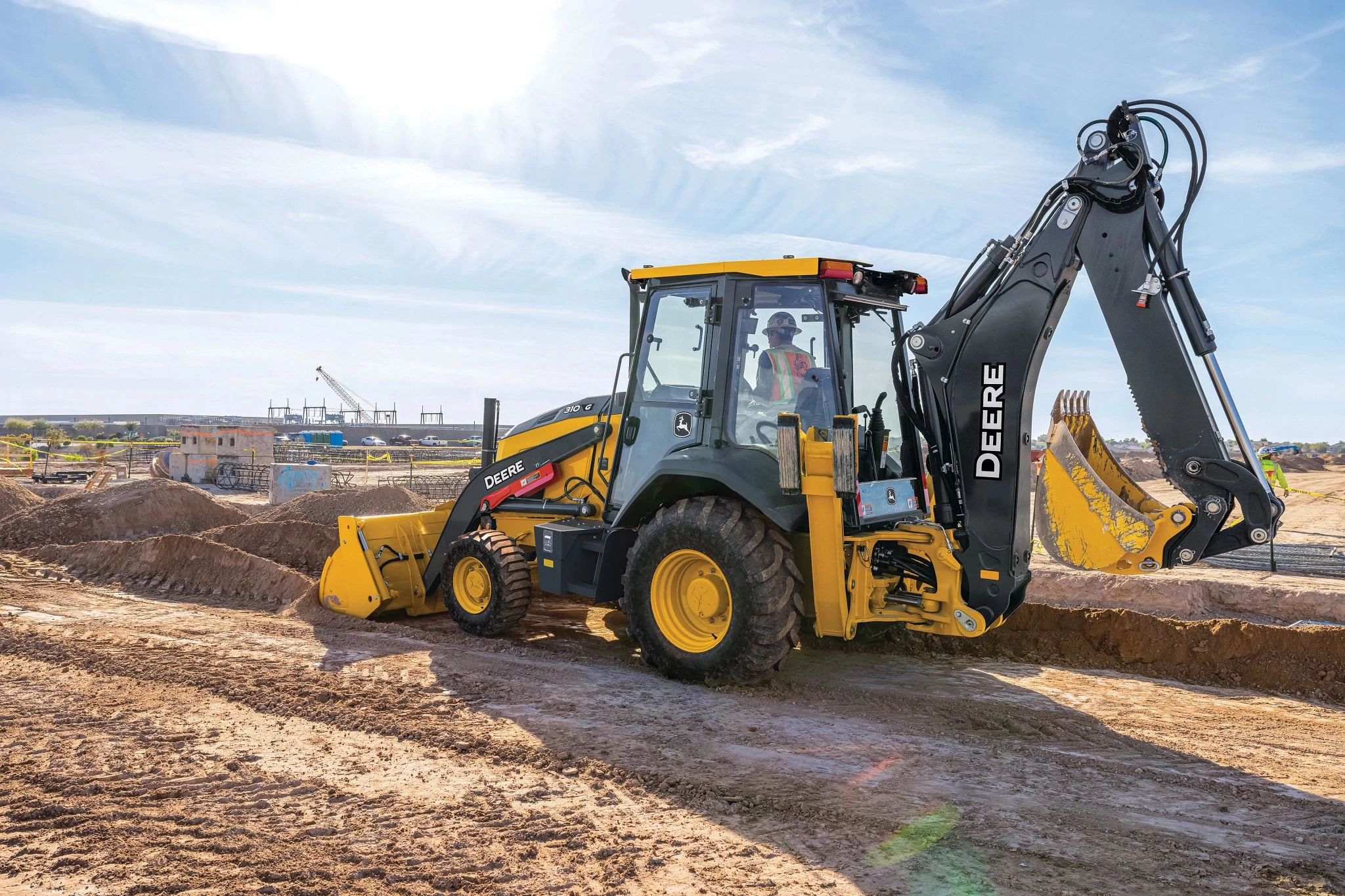 A yellow John Deere backhoe loader operates at an active construction site, positioned on sandy, uneven ground.