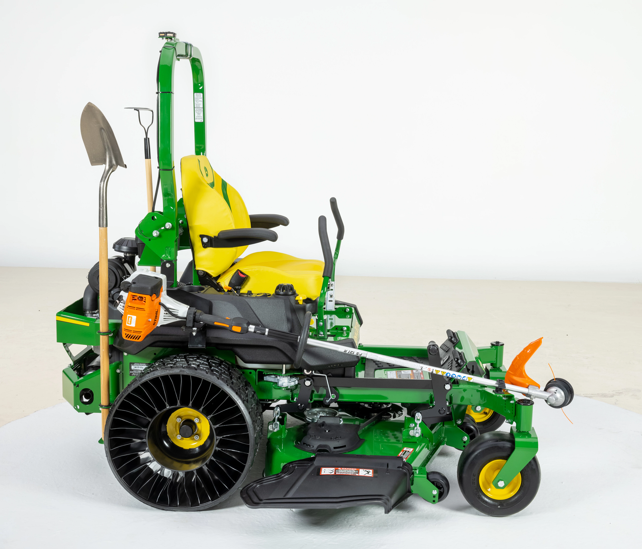 Side view of John Deere zero-turn mower featuring a roll bar, large rear wheels, front caster wheels, and mounted landscaping tools including a shovel and a string trimmer.