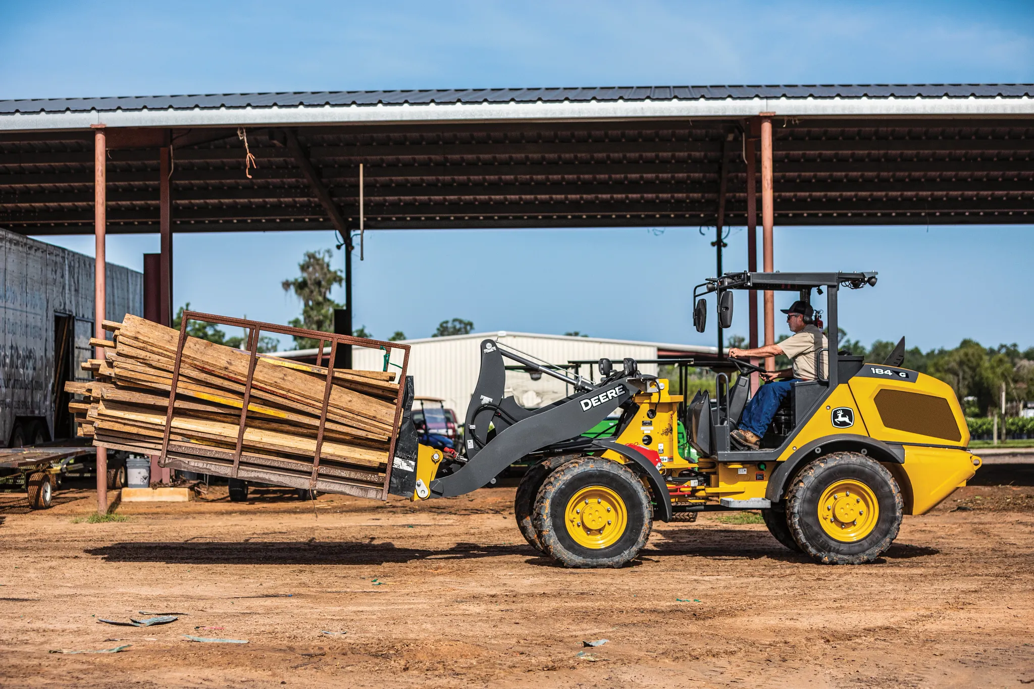 A wheel loader using pallet forks to carry a stacked bundle of lumber across a dirt yard, with an open-sided metal structure and additional materials visible in the background.