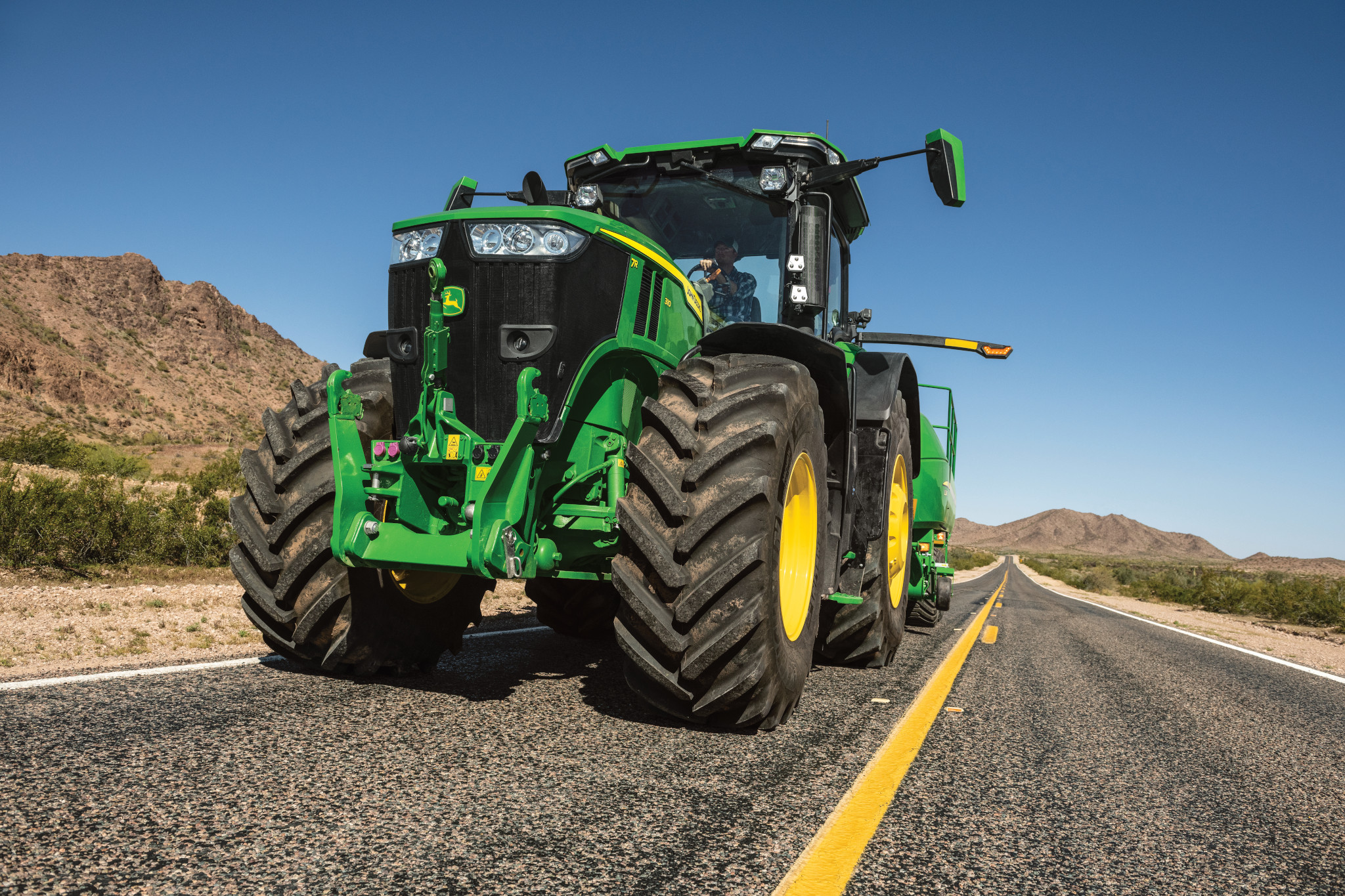 Green tractor driving down an empty paved road with a yellow centerline, with large tires and detailed front end shown against a clear blue sky, with dry, rocky hills and sparse vegetation in the background