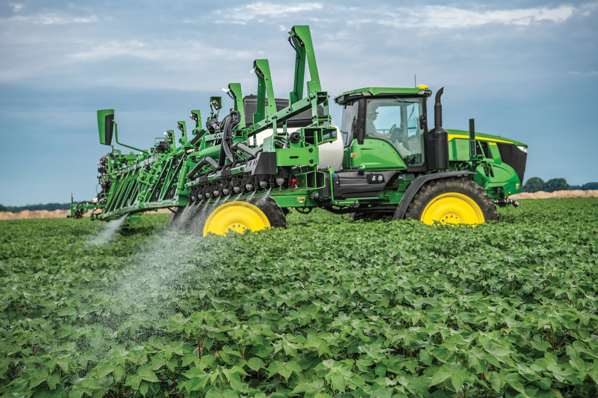 Large green agricultural sprayer with wide extended booms applying liquid across a dense, leafy crop field, with fine mist spraying onto the plants under an overcast sky