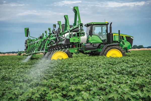 Large green agricultural sprayer with wide extended booms applying liquid across a dense, leafy crop field, with fine mist spraying onto the plants under an overcast sky