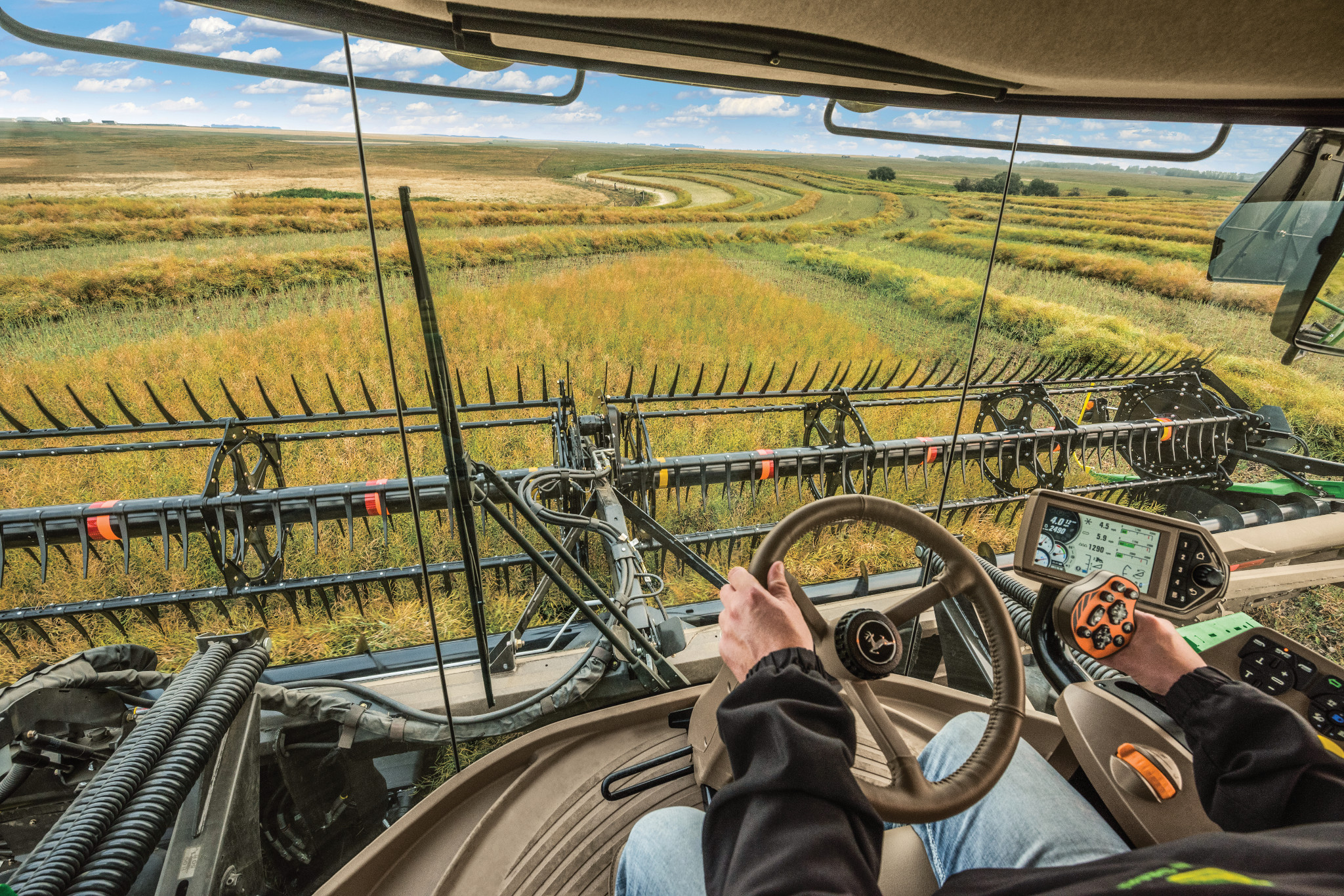 Operator’s view from inside a John Deere self‑propelled windrower cab, hands on steering wheel and controls, looking over wide draper header cutting hay into curved windrows across an open field.
