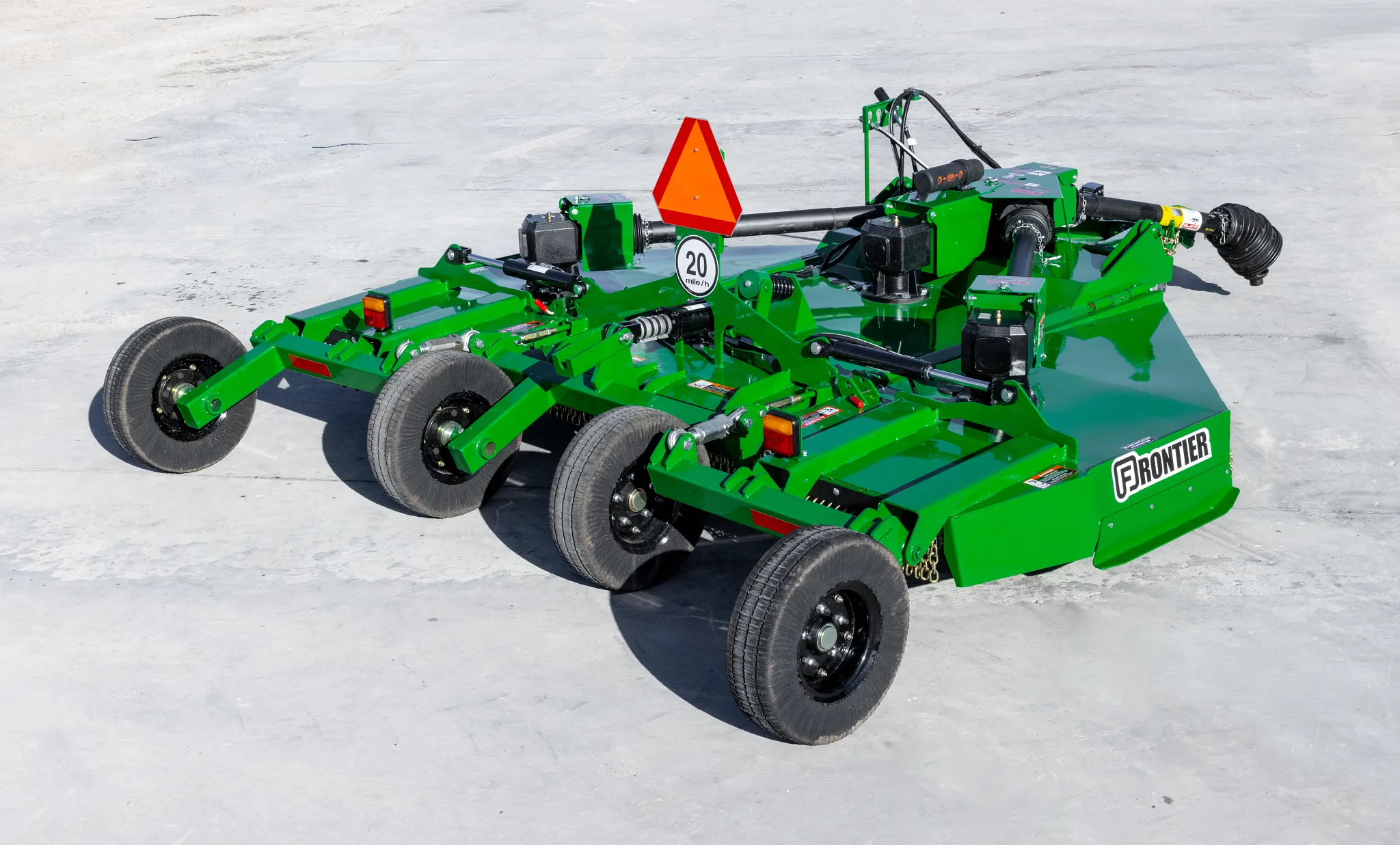 Rear three-quarter view of a green Frontier rotary cutter with four transport wheels, PTO driveline, hydraulic components, safety chain shielding, and a slow-moving vehicle emblem, parked on a concrete surface outdoors