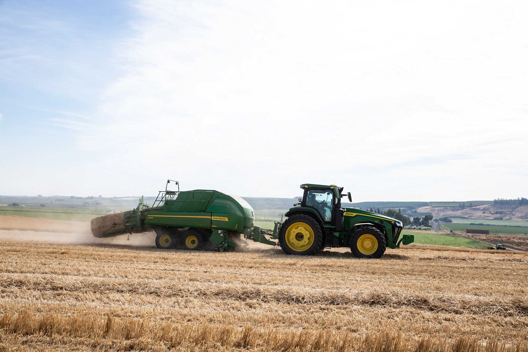 John Deere tractor pulling a baler across a harvested field, producing dust as it operates in an open rural landscape
