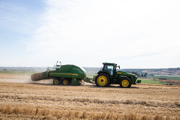 John Deere tractor pulling a baler across a harvested field, producing dust as it operates in an open rural landscape