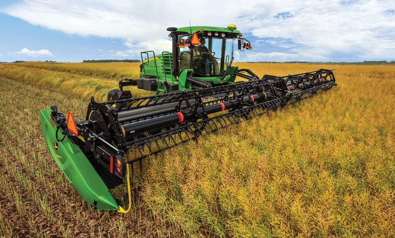 A self-propelled windrower with a wide draper header harvests a golden crop in a field beneath a cloudy sky.