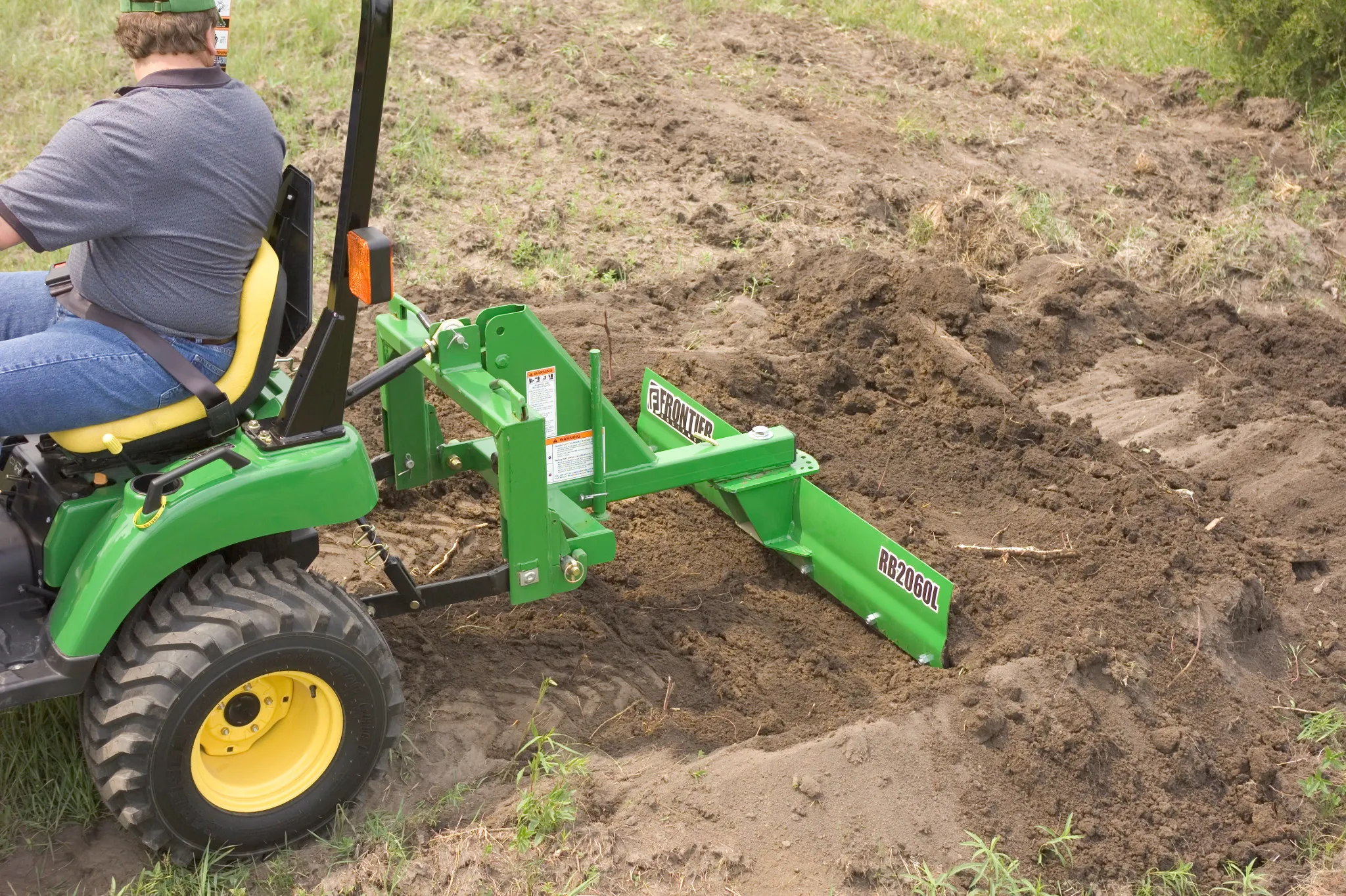 Side view of a green RB2060L Rear Blade at work in dirt, with half of the attached tractor visible and operator atop it turning back to look at the blade.