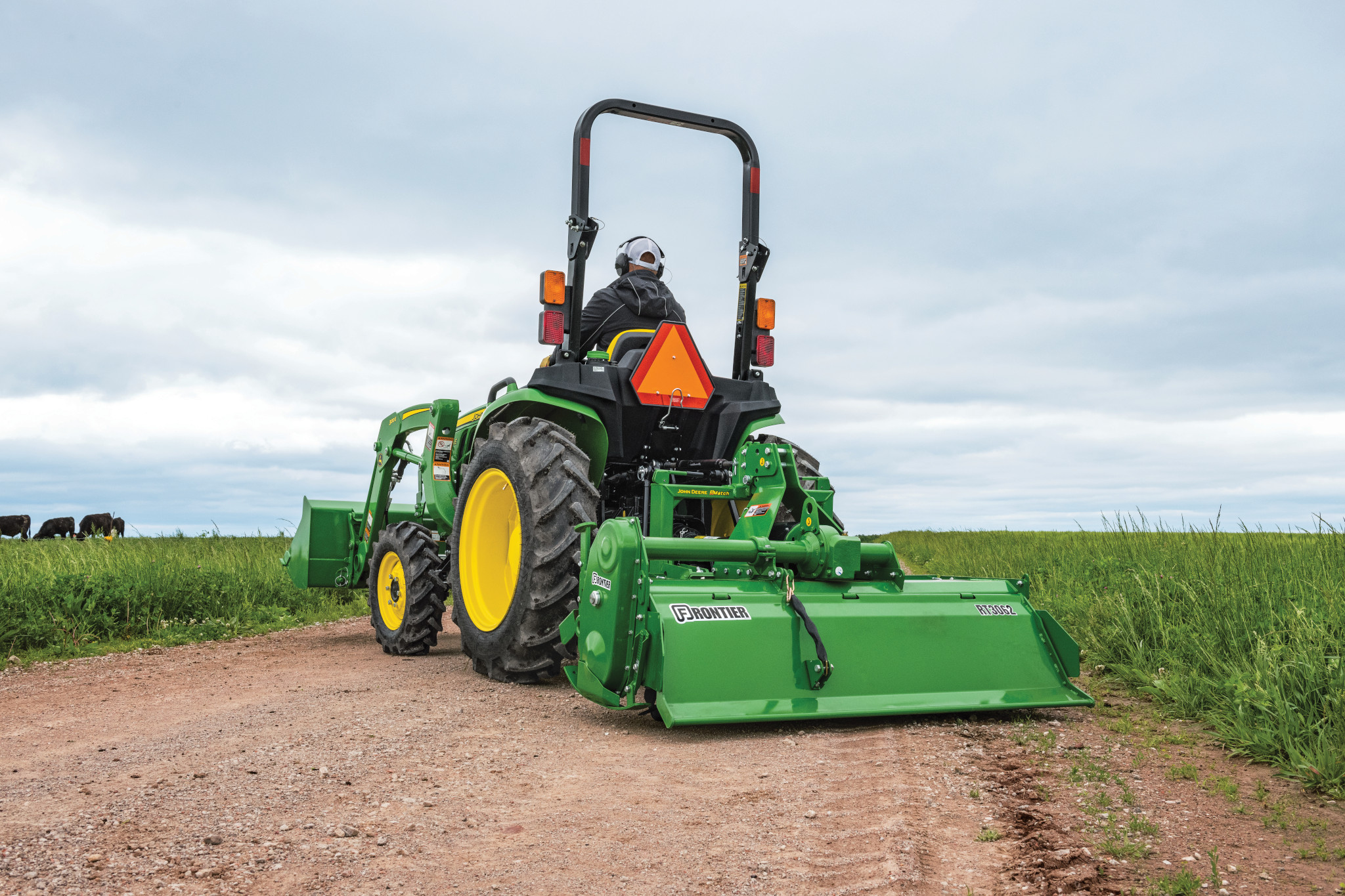 Rear view of a compact tractor driving on a dirt path with a rotary tiller attached, working along a grassy field under a cloudy sky