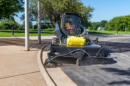 Front view of a BP84C pick-up broom with optional roof-mounted water tank cleaning the street along a curb on the front of a skid steer