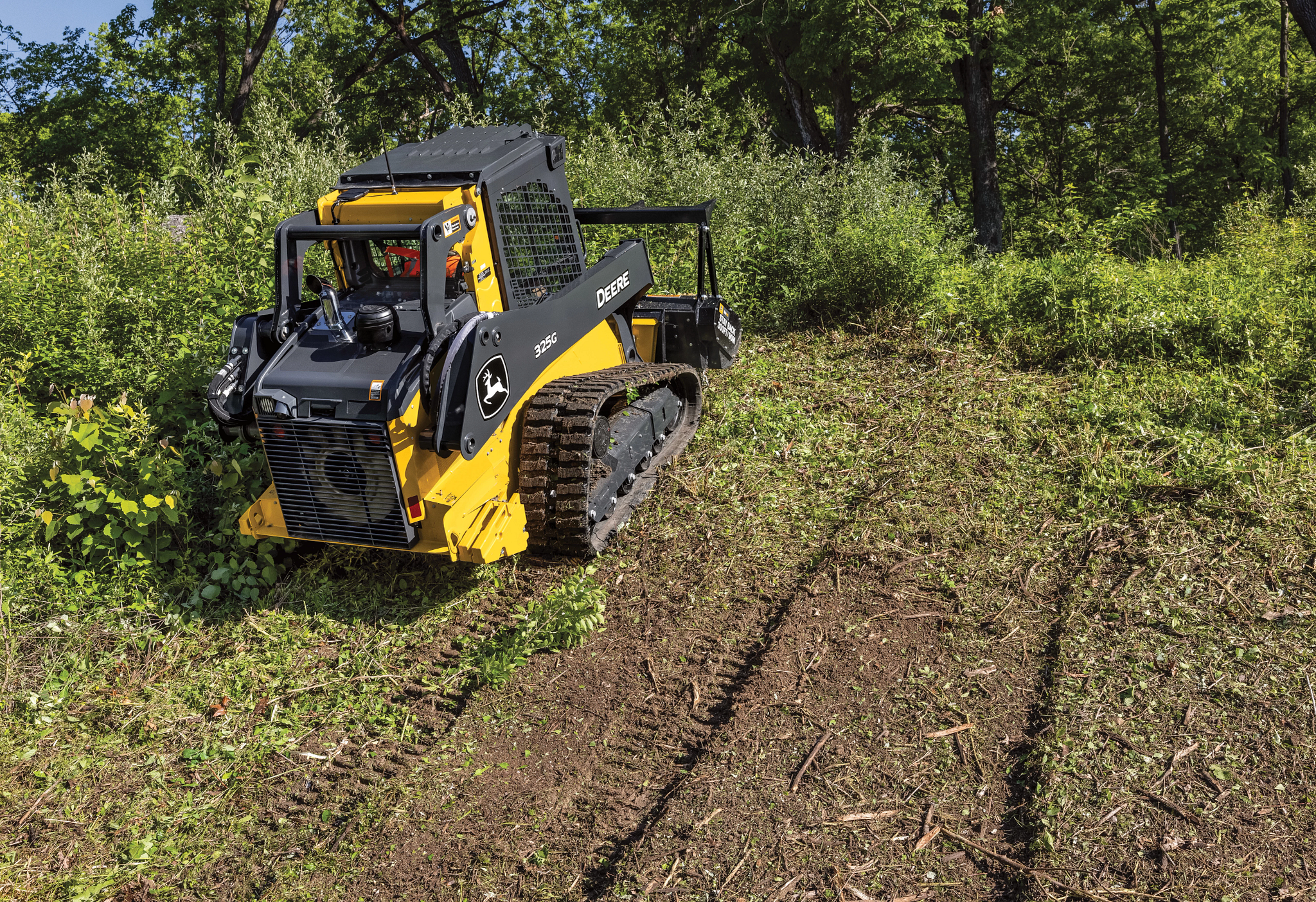 John Deere compact track loader operating on a cleared dirt path in a wooded area, featuring a yellow and gray body