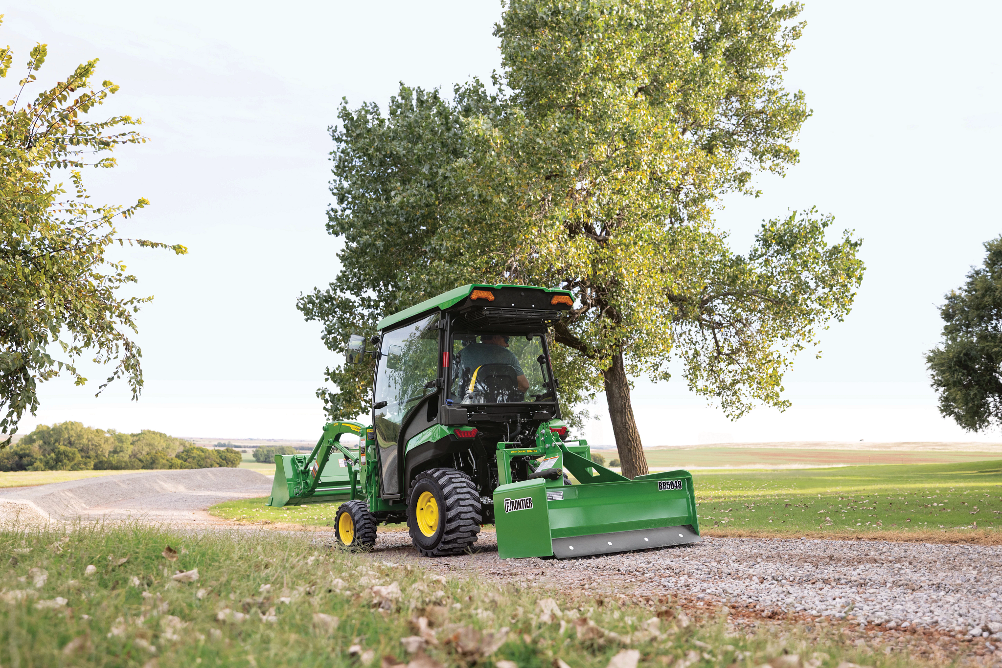A green tractor with an enclosed cab pulls a box blade attachment along a gravel path beside a tree, with grassy fields and a flat rural landscape under a clear sky